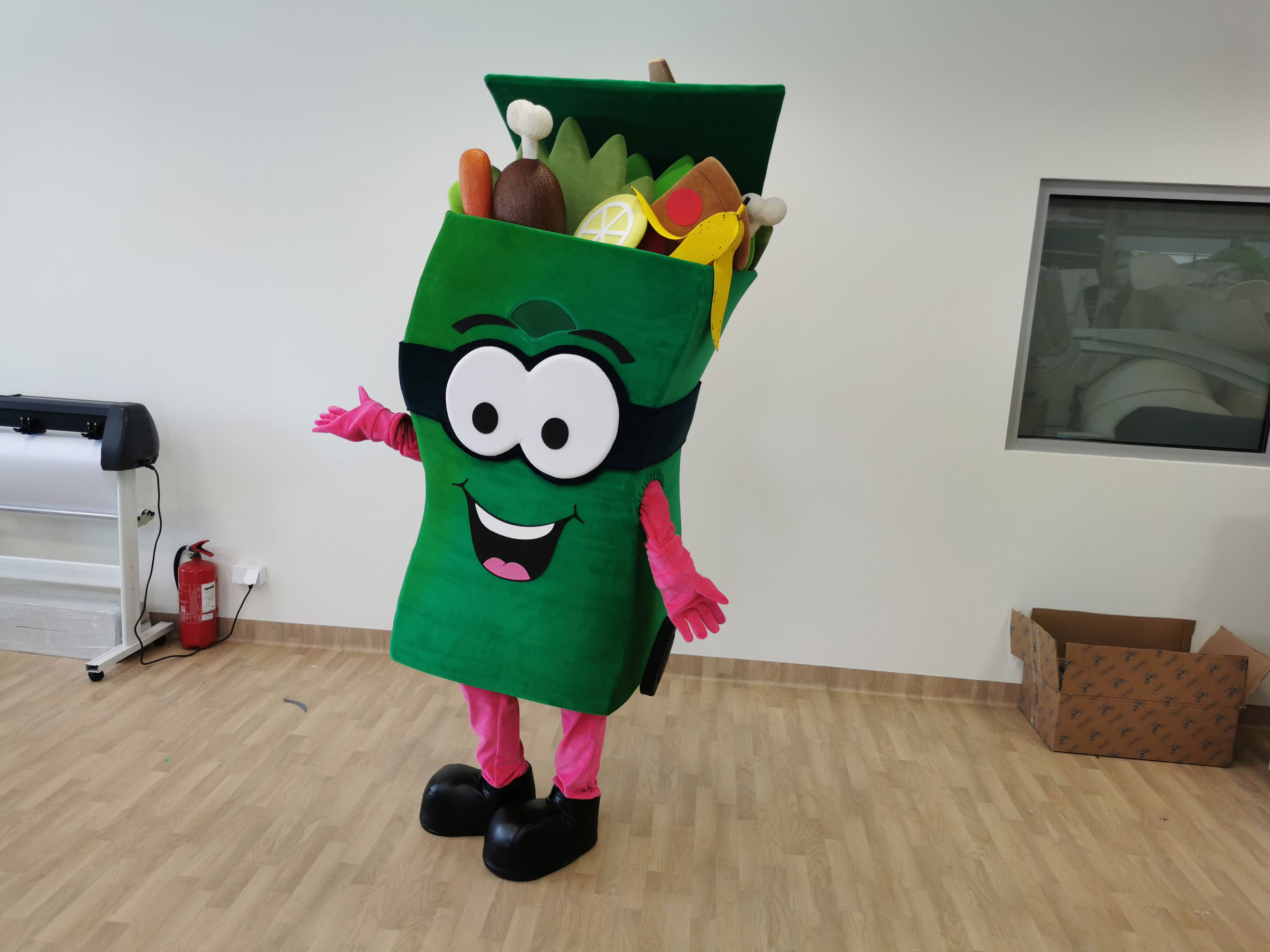 Na fotografii widnieje maskotka reklamowa w postaci zielonego kontenera na śmieci z widocznymi spod klapy różnorodnymi odpadkami, pozujący do zdjęcia w siedzibie firmy Elabika